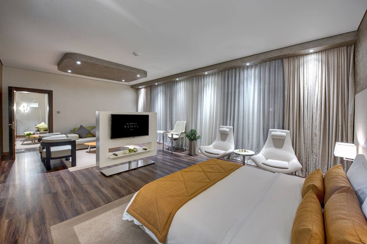 A modern hotel room with a large bed, a flat-screen TV, a seating area with chairs, and a dining table.