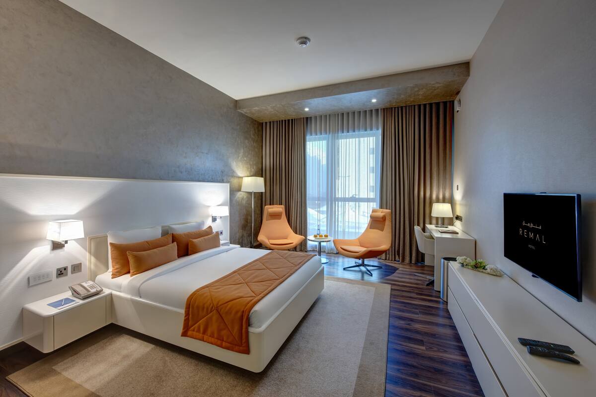 A modern hotel room with a large bed, two armchairs, a TV, and a small table.