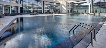 Uma piscina interior moderna com paredes de vidro transparente, espreguiçadeiras e uma escada de metal.