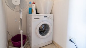 Un área de lavandería compacta con lavadora de carga frontal, un ventilador de pie y un cubo morado.