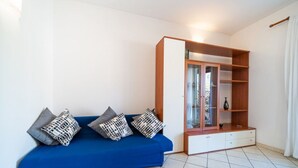 Una habitación con un sofá azul, cojines estampados, un aparador de madera con puertas de cristal y una cortina blanca.