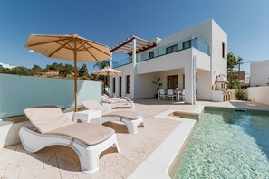 Un área moderna junto a la piscina con sillones de descanso y sombrillas, una piscina cristalina y un edificio contemporáneo con balcón.