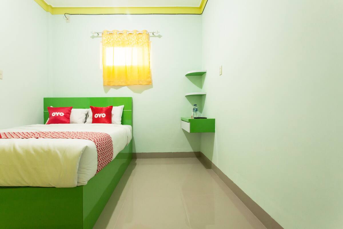 Une chambre à coucher avec une tête de lit verte, un lit avec une literie rouge et blanche, une petite table de chevet verte avec une lampe, et une fenêtre avec un rideau jaune.