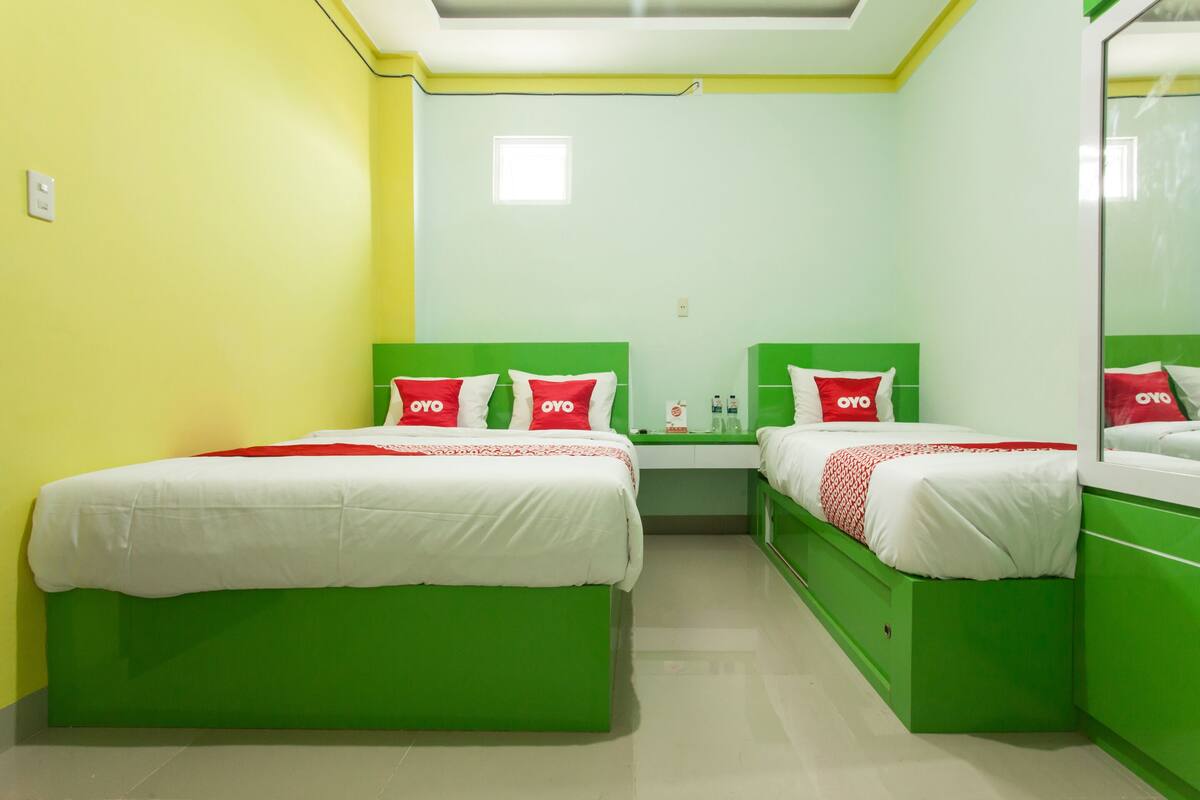 Une chambre avec deux lits, chacun doté d’une tête de lit verte et de draps blancs. On y trouve une petite table avec un miroir, ainsi qu’un coussin rouge et blanc placé entre les deux lits.