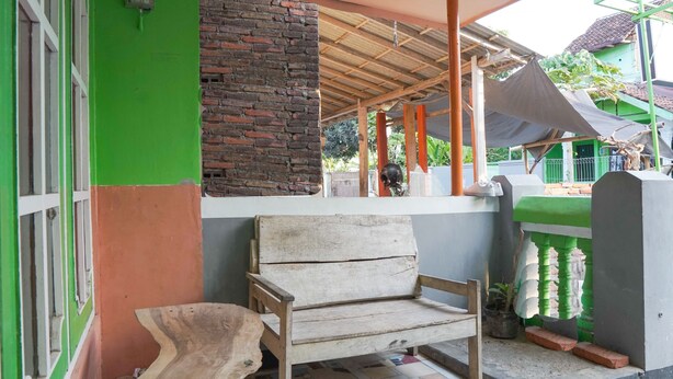 A covered outdoor seating area with a wooden bench, a brick wall, and a green and orange building facade.