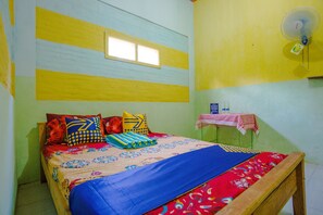 A bed with colorful bedding, a window, a fan, and a small table with a checkered tablecloth.