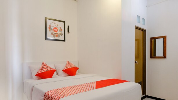 A neatly made bed with a patterned bedspread and two red pillows, a framed floral artwork on the wall, and a door with a mirror on the adjacent wall. - Hotel O Kina Family Residence Syariah (Malang)