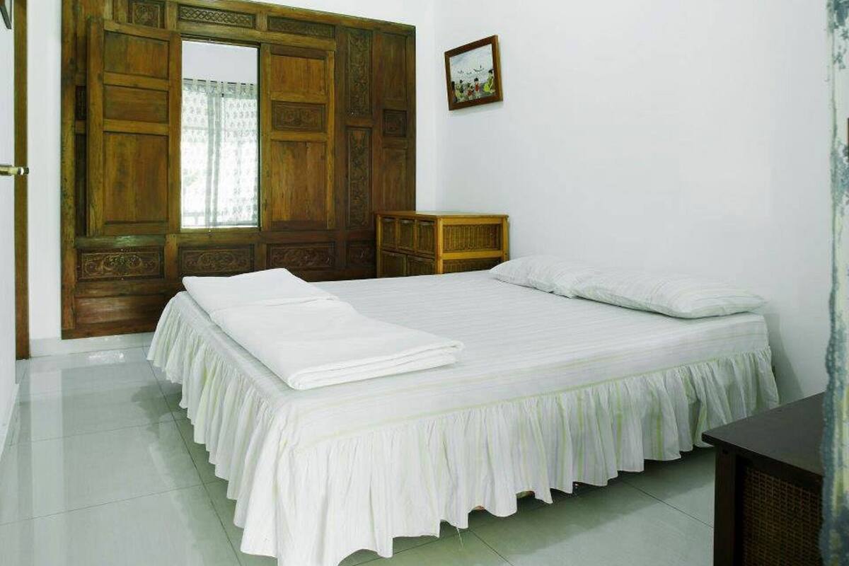 A bedroom with a large wooden wardrobe, a bed with white bedding, a small wooden nightstand, and a framed picture on the wall.