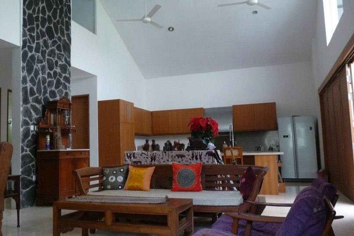 A spacious living room with a high ceiling, wooden furniture, and a kitchen area with wooden cabinets and a refrigerator.
