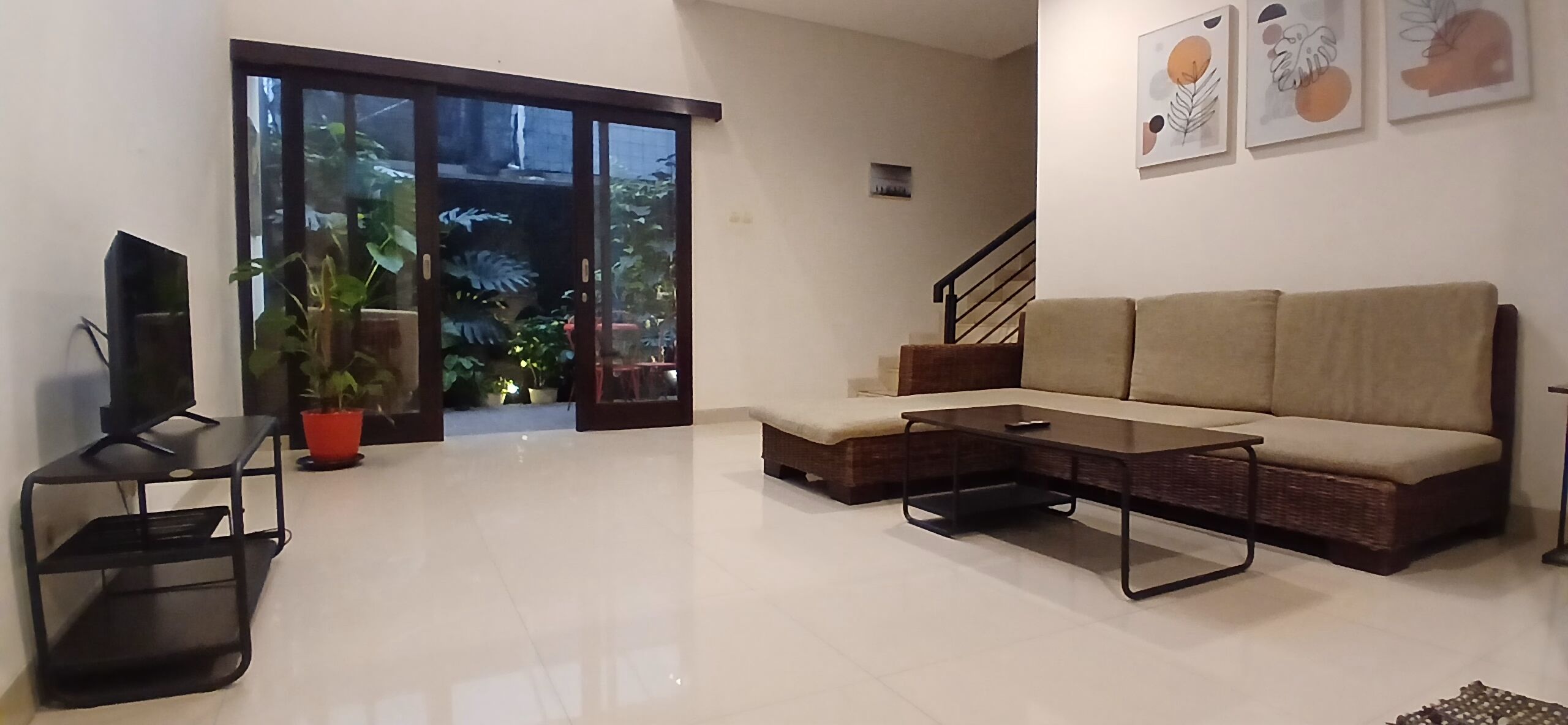 Una sala de estar con un sofá, una mesa de centro, un televisor sobre un mueble bajo y un ventanal corredizo que da a un balcón con plantas.