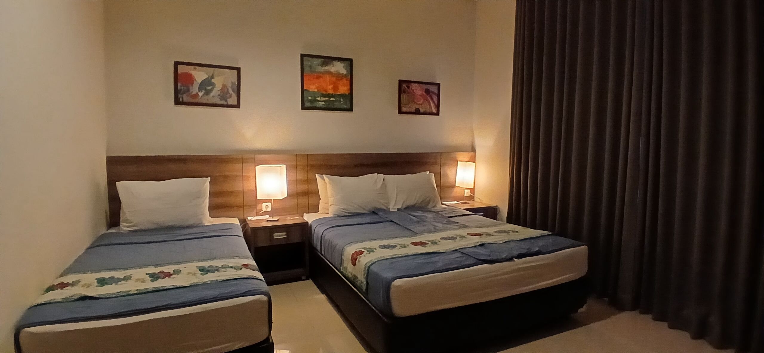 A hotel room with two beds, each with a wooden headboard, bedside lamps, and three framed artworks on the wall.