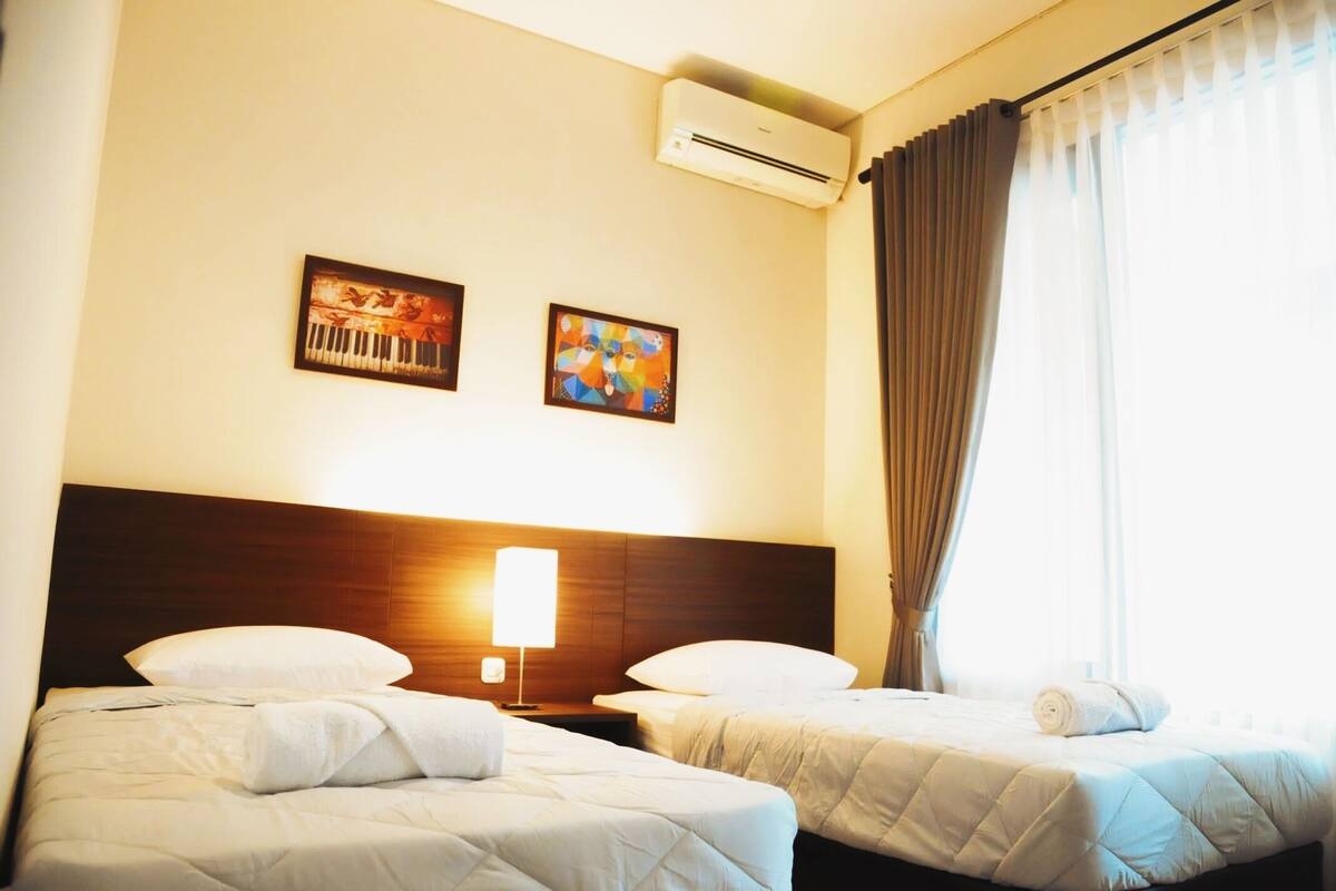 Une chambre d’hôtel avec deux lits, une tête de lit, un appareil de climatisation et deux tableaux accrochés au mur.