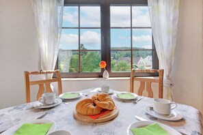 A dining table set with plates, a croissant, a vase with a flower, and a window with a view of houses and trees.