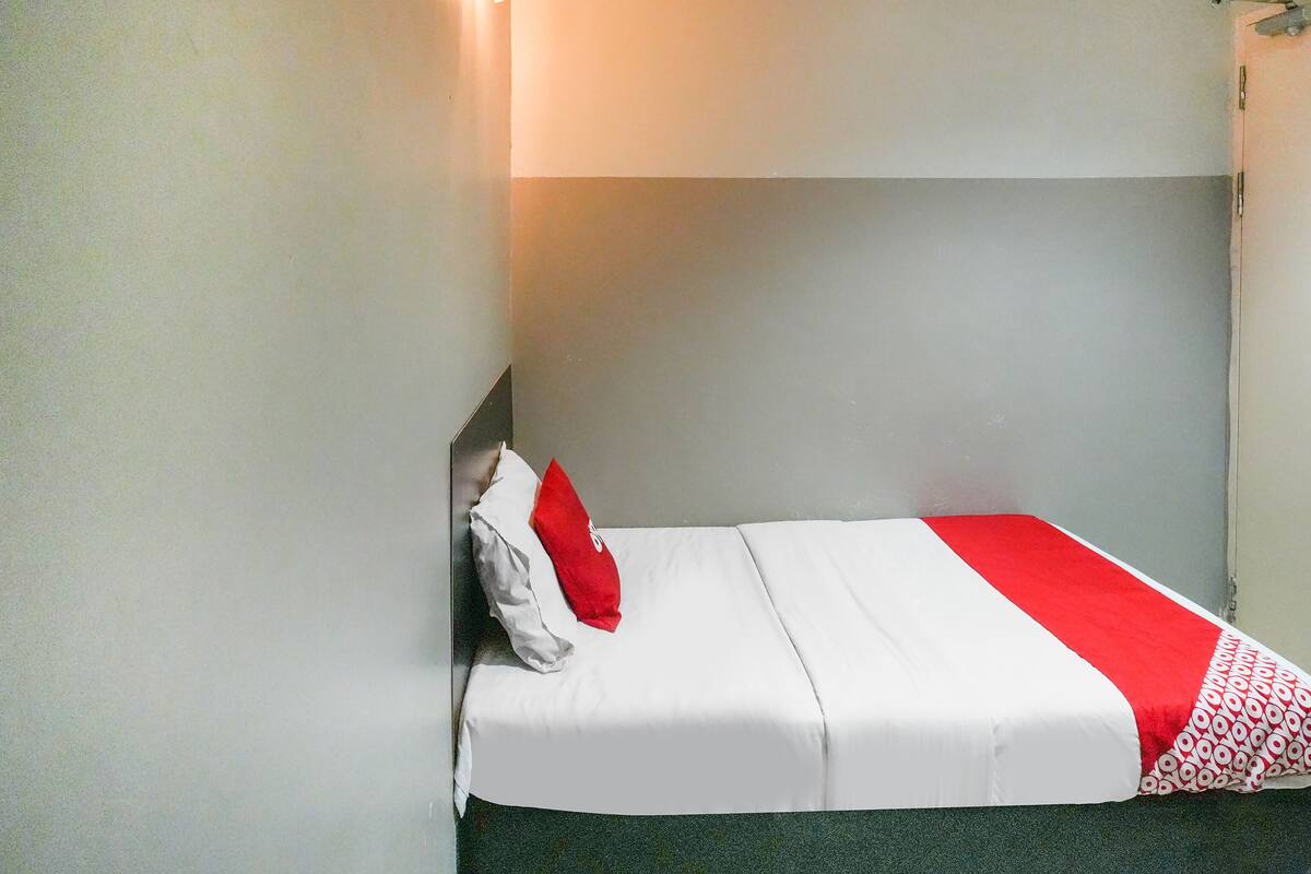 Un llit petit i ben fet amb roba de llit blanca i vermella en una habitació amb parets lises i una sola làmpada.