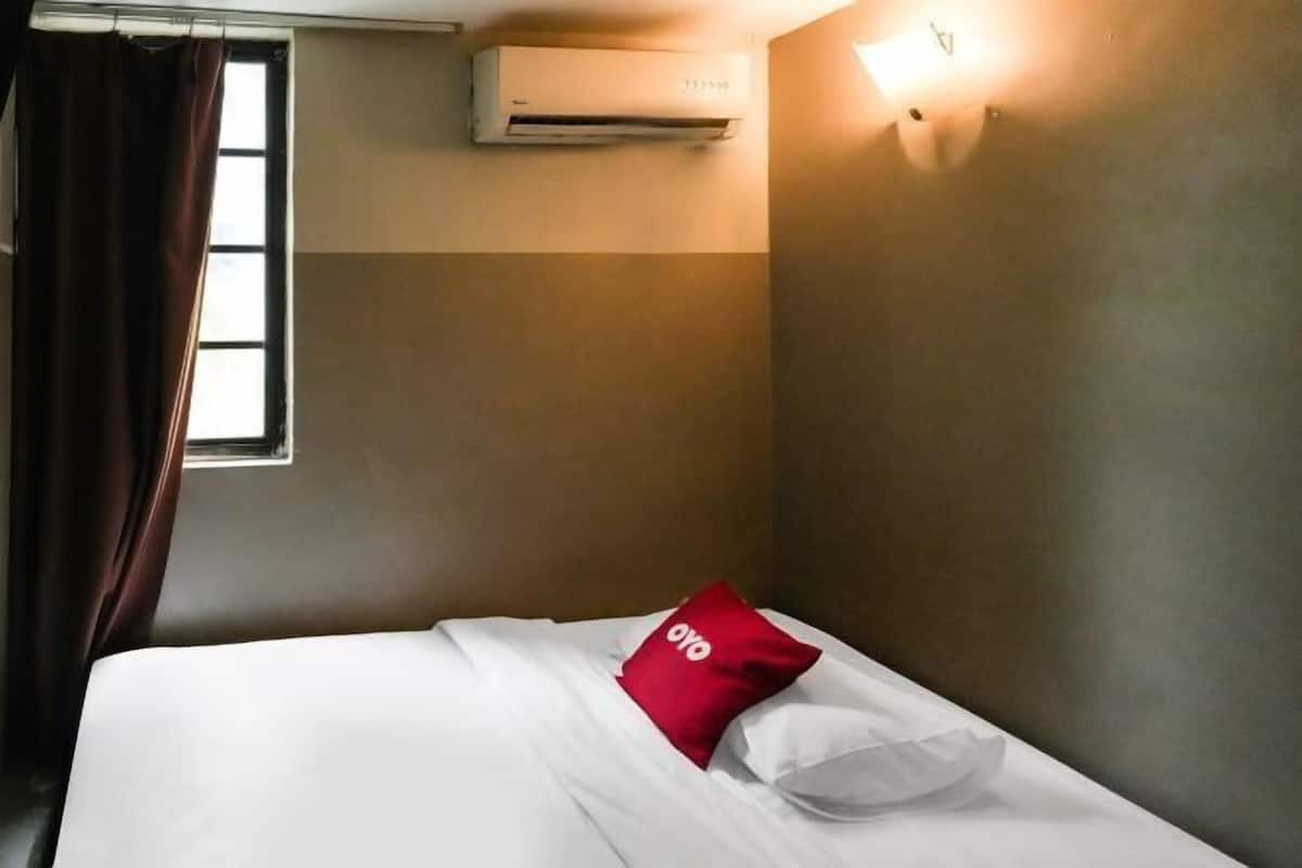 Habitació d'hotel amb llit, finestra amb cortines, aparell d'aire condicionat i llum penjant a la paret.