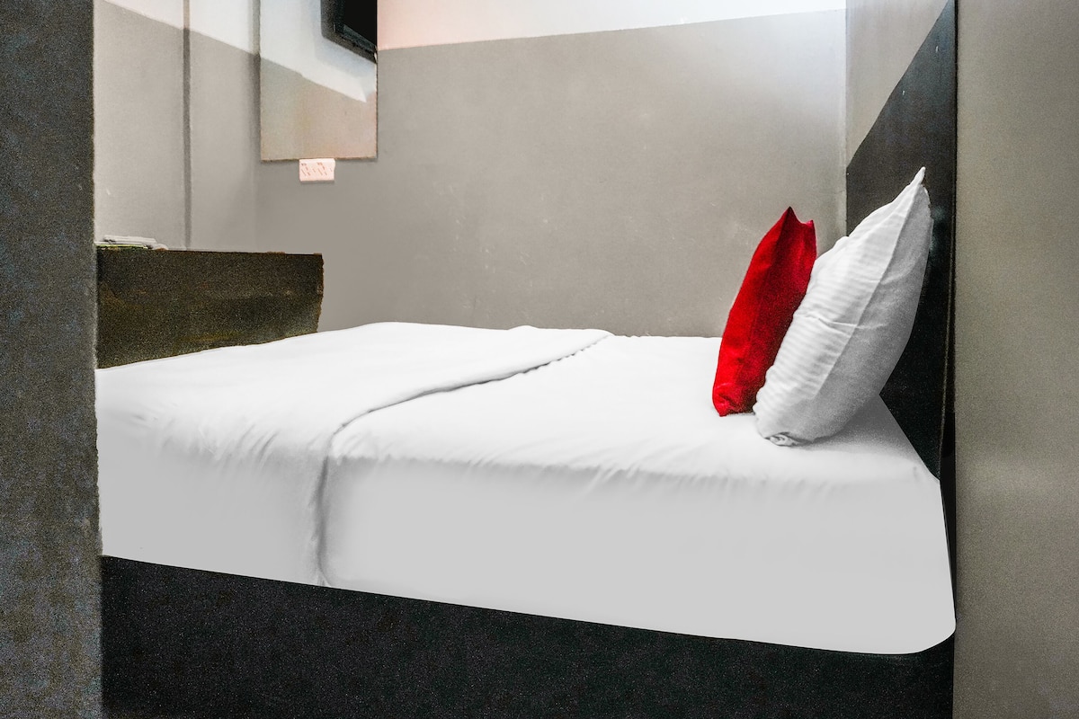 Habitació d'hotel moderna amb un llit, roba de llit blanca, un coixí vermell i un mirall a la paret.