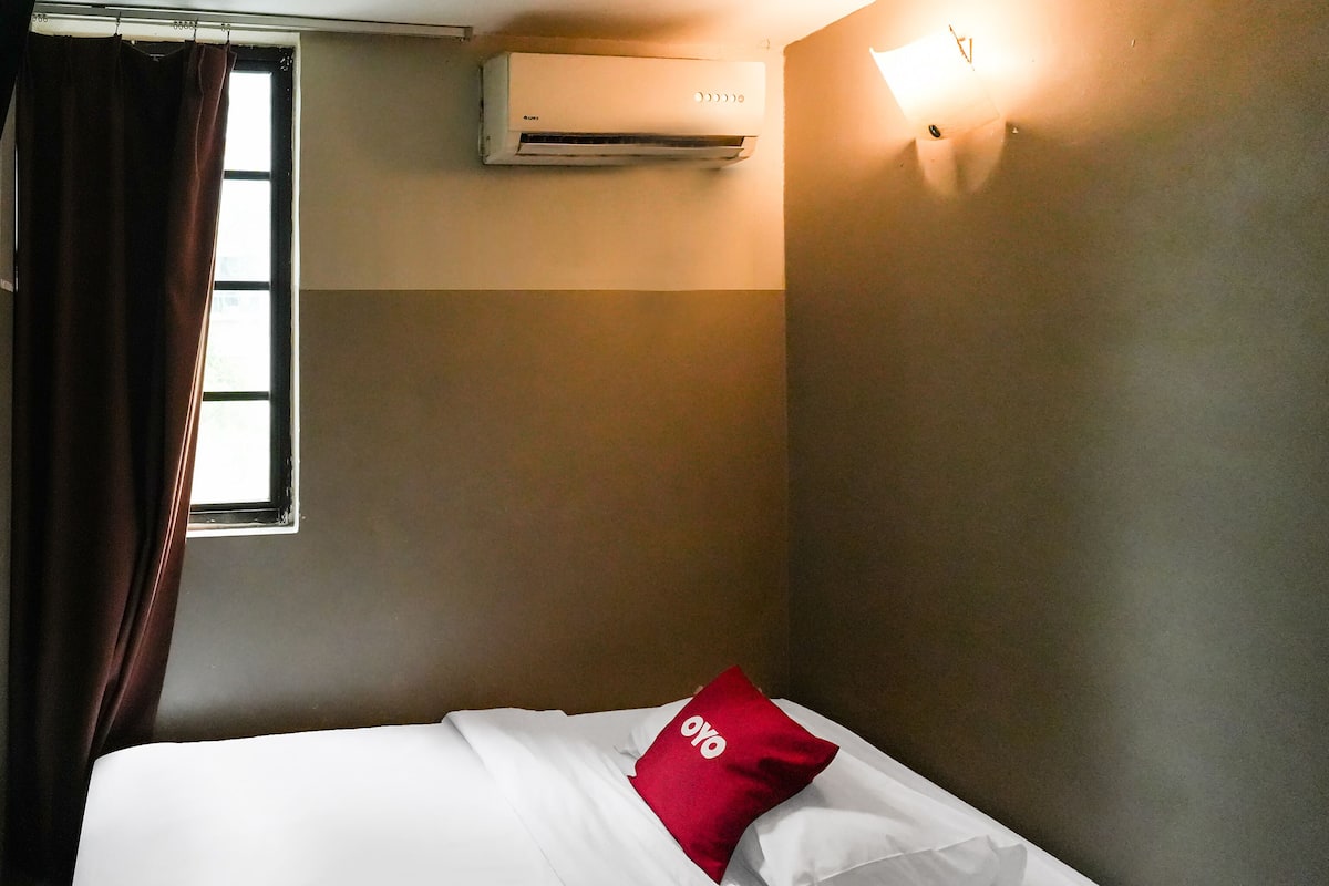 Habitació d'hotel amb un llit, una finestra amb cortines, un aparell d'aire condicionat i una làmpada fixada a la paret.