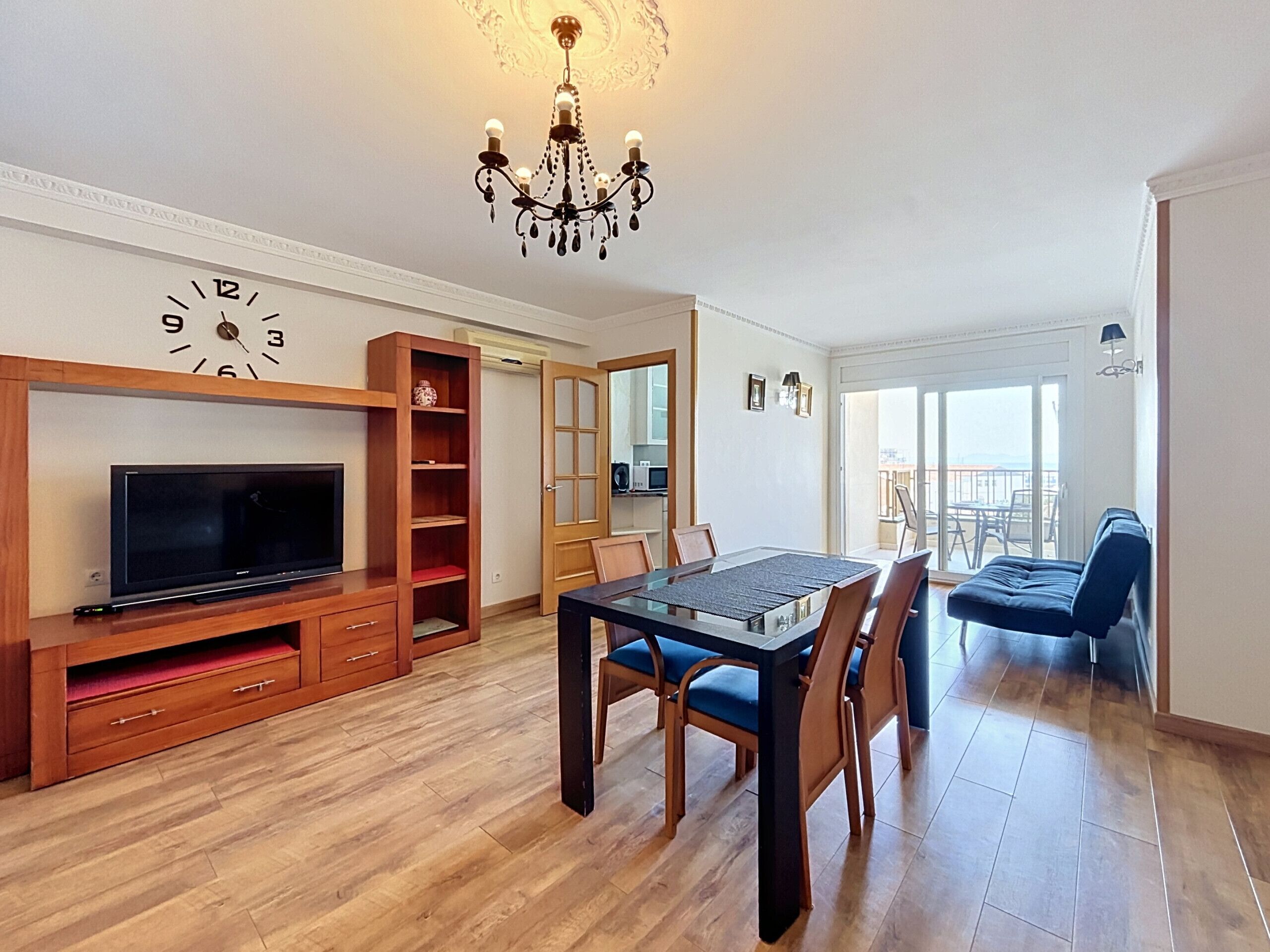 A modern living room with a flat-screen TV, wooden entertainment unit, dining table with chairs, and a chandelier.