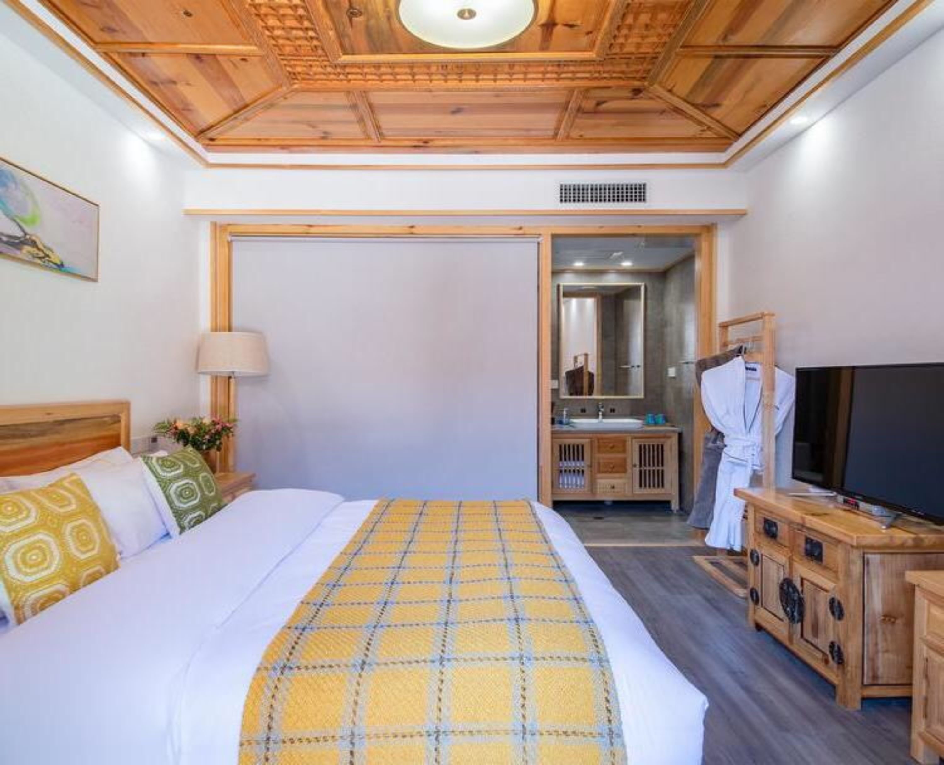A bedroom with a wooden ceiling, a bed with a yellow and white checkered blanket, a TV on a wooden cabinet, and a bathroom visible through an open door.