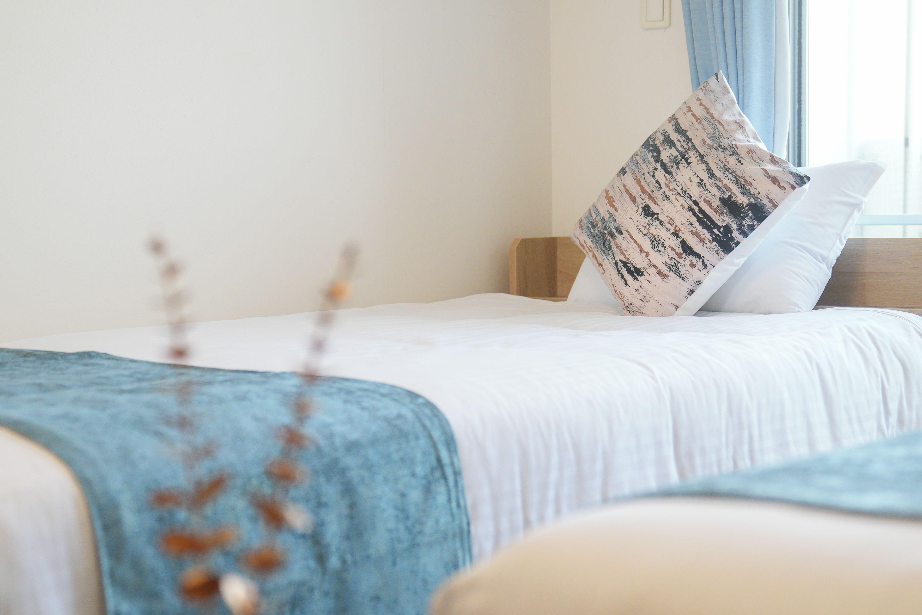 A neatly made bed with a white duvet and a patterned pillow, a wooden headboard, and a window with curtains.