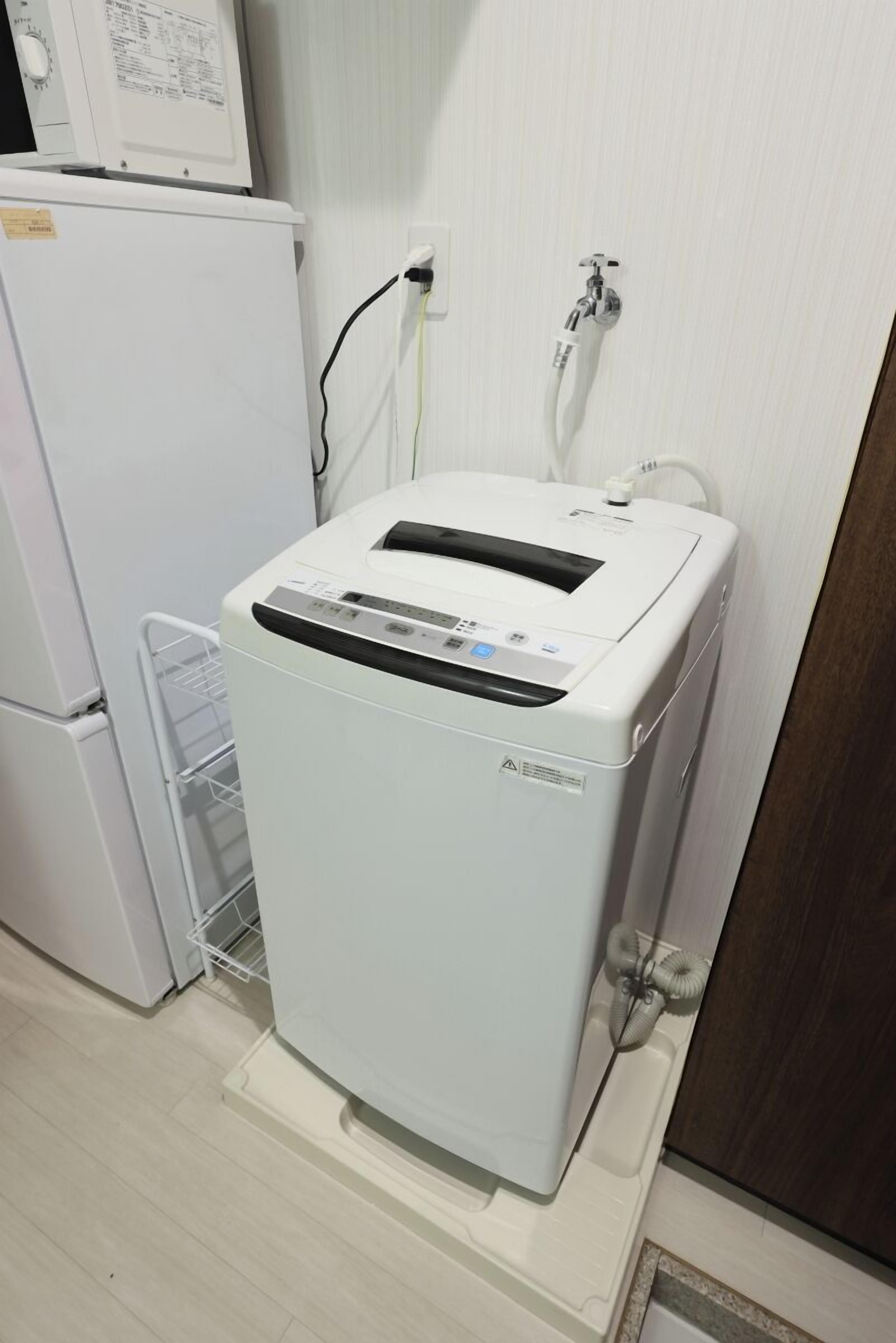 A compact, white, wall-mounted laundry machine with a control panel, located in a small, clean room with a white refrigerator and a white shelf.