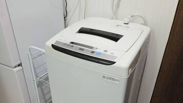 A compact, white, wall-mounted laundry machine with a control panel, located in a small, clean room with a white refrigerator and a white shelf.