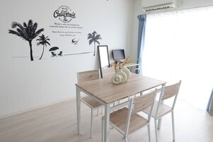 Un comedor con mesa y sillas de madera, un adhesivo mural con palmeras y texto, un cuadro enmarco y una mesita con adornos.
