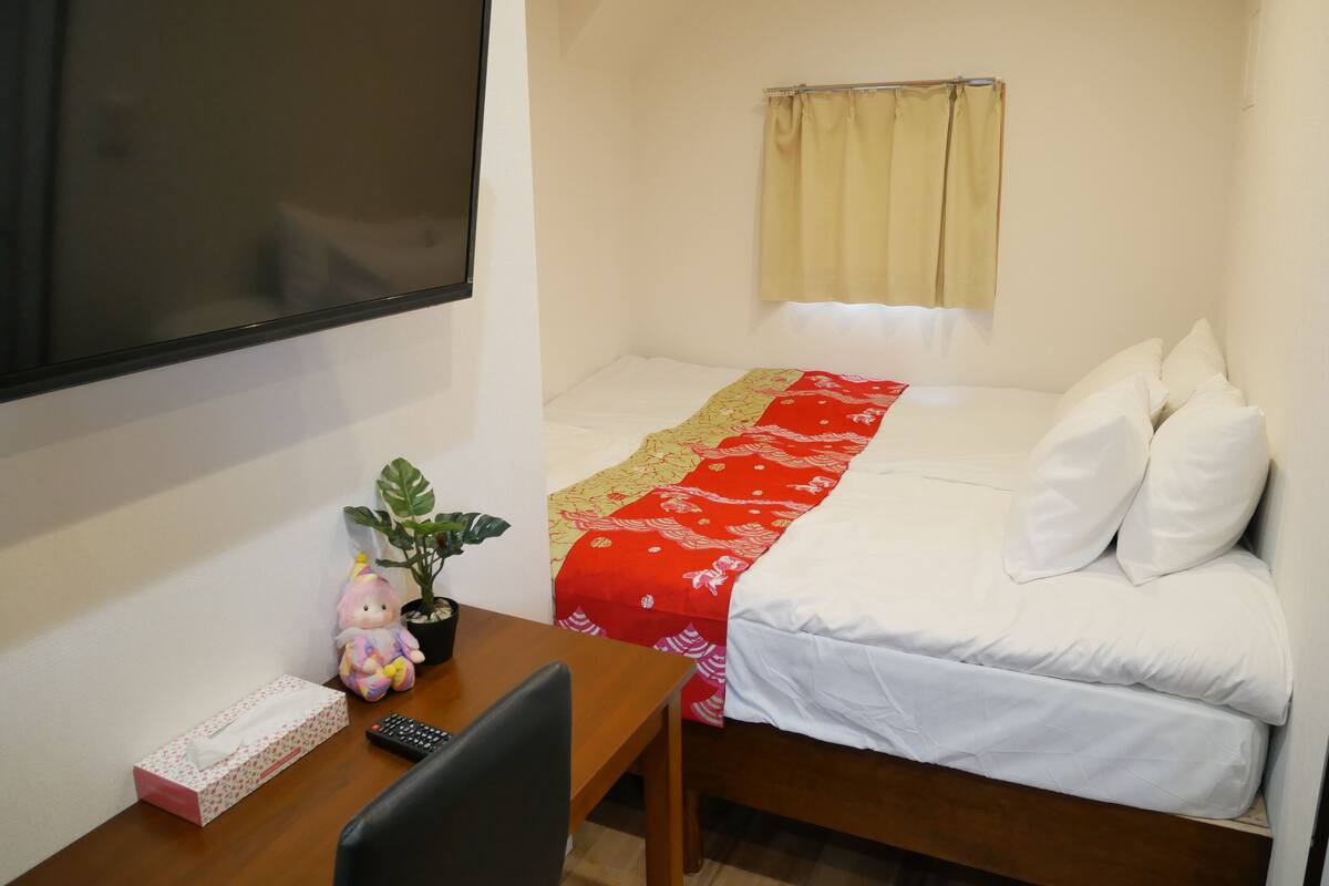 Une petite chambre à coucher, bien rangée, avec un lit, un bureau, une télévision et une fenêtre avec des rideaux.