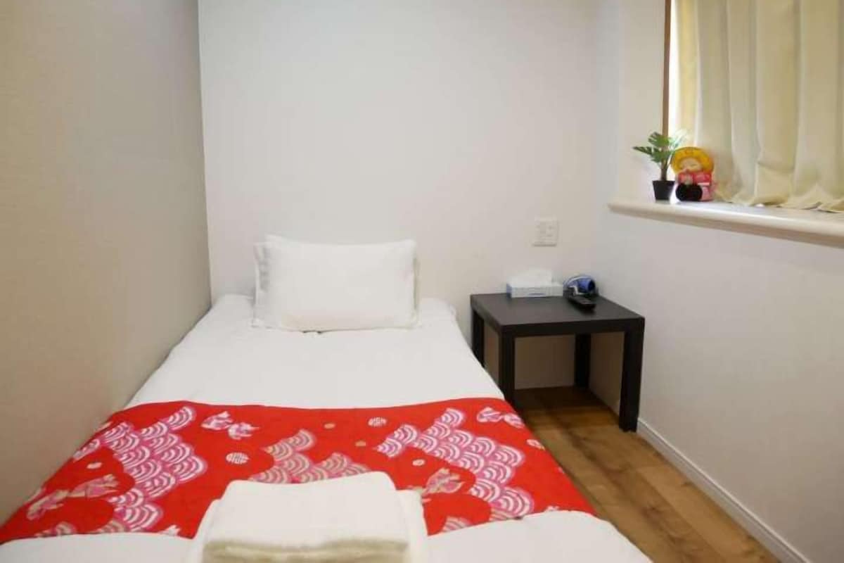Ein kleines Zimmer mit einem Einzelbett, einer rot gemusterten Tagesdecke, einem weißen Kissen, einem kleinen Nachttisch mit Lampe und einem Fenster mit Vorhängen.
