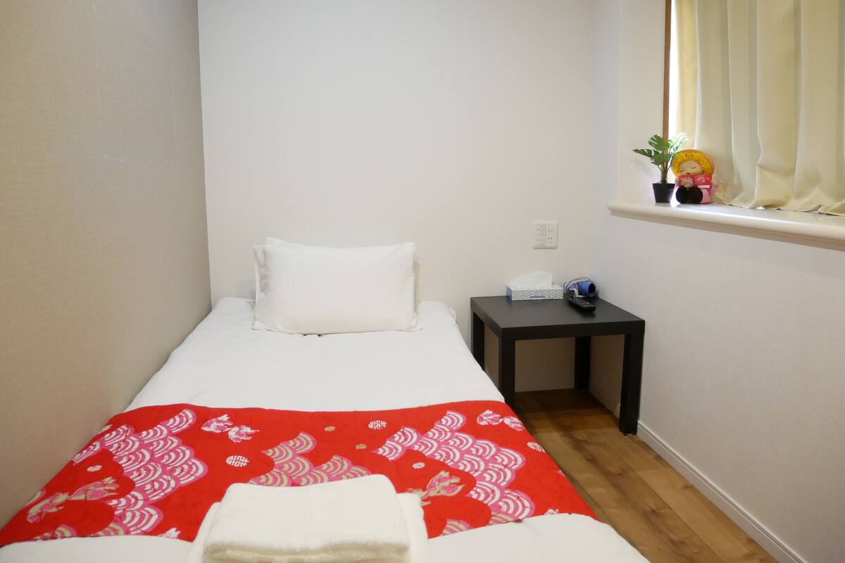 Une petite chambre avec un lit simple, recouverte d’une courtepointe à motifs rouges, un oreiller blanc, une petite table avec une télécommande, une boîte de mouchoirs et une plante en pot près de la fenêtre.