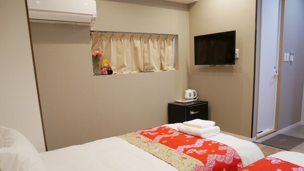 Ein Hotelzimmer mit zwei Betten, einem kleinen Nachttisch mit Wasserkocher, einem an der Wand montierten Fernseher und einem Fenster mit Vorhängen.