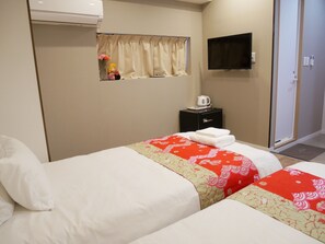 Quarto de hotel com duas camas, uma pequena mesa de cabeceira com chaleira, televisão na parede e janela com cortinas.