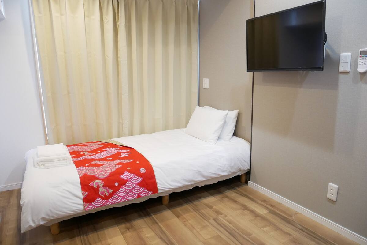 Un lit simple avec une couvre-lit rouge et blanche, un téléviseur fixé au mur et un plancher en bois.