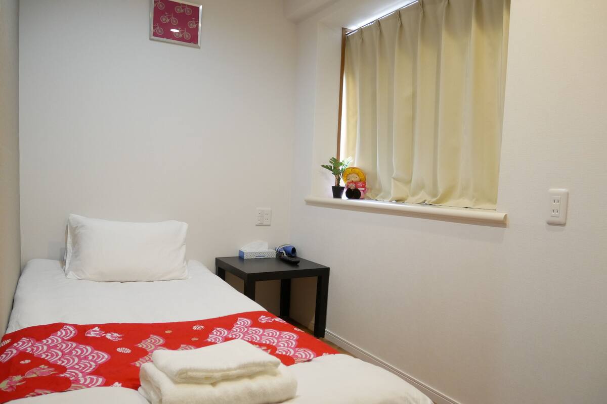 Un petit lit bien fait, avec des draps blancs et une couverture rouge à motifs. Une petite table se trouve à côté du lit, sur laquelle reposent une boîte de mouchoirs et une télécommande. On aperçoit une fenêtre avec un rideau et une plante en pot.