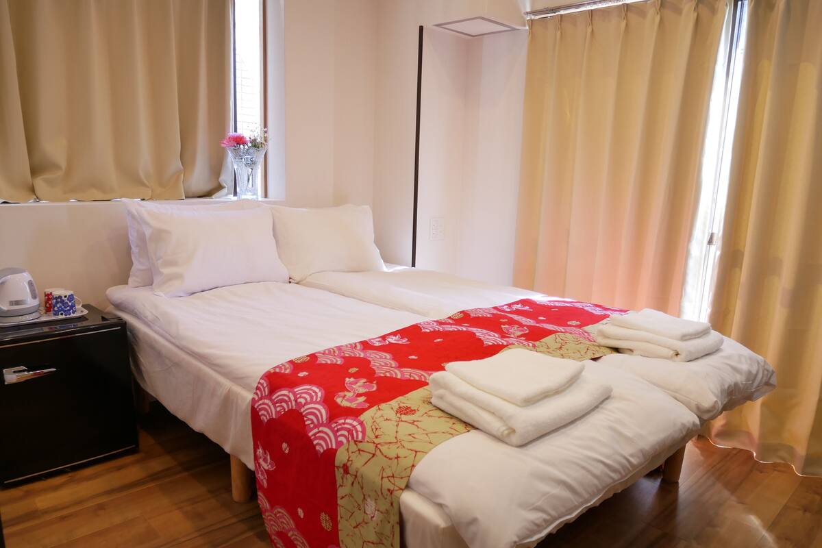 Un lit bien fait, avec du linge de lit blanc, une couverture à motifs rouges et blancs, et une table de chevet sur laquelle se trouvent une bouilloire et une tasse.