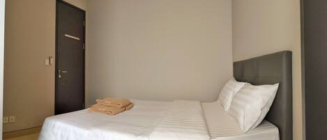 Una cama bien tendida con sábanas blancas y dos toallas color beige, un cabecero oscuro y una pared lisa con una puerta.
