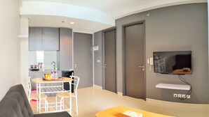 A modern hotel room with a kitchen area, a dining table with chairs, a flat-screen TV mounted on the wall, and a small shelf below the TV. -  Lovina at One Residence (Batam)