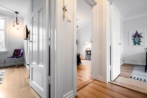 Un couloir doté d’un plancher en bois, de murs blancs et d’une porte donnant sur une pièce où se trouvent une chaise et une table.