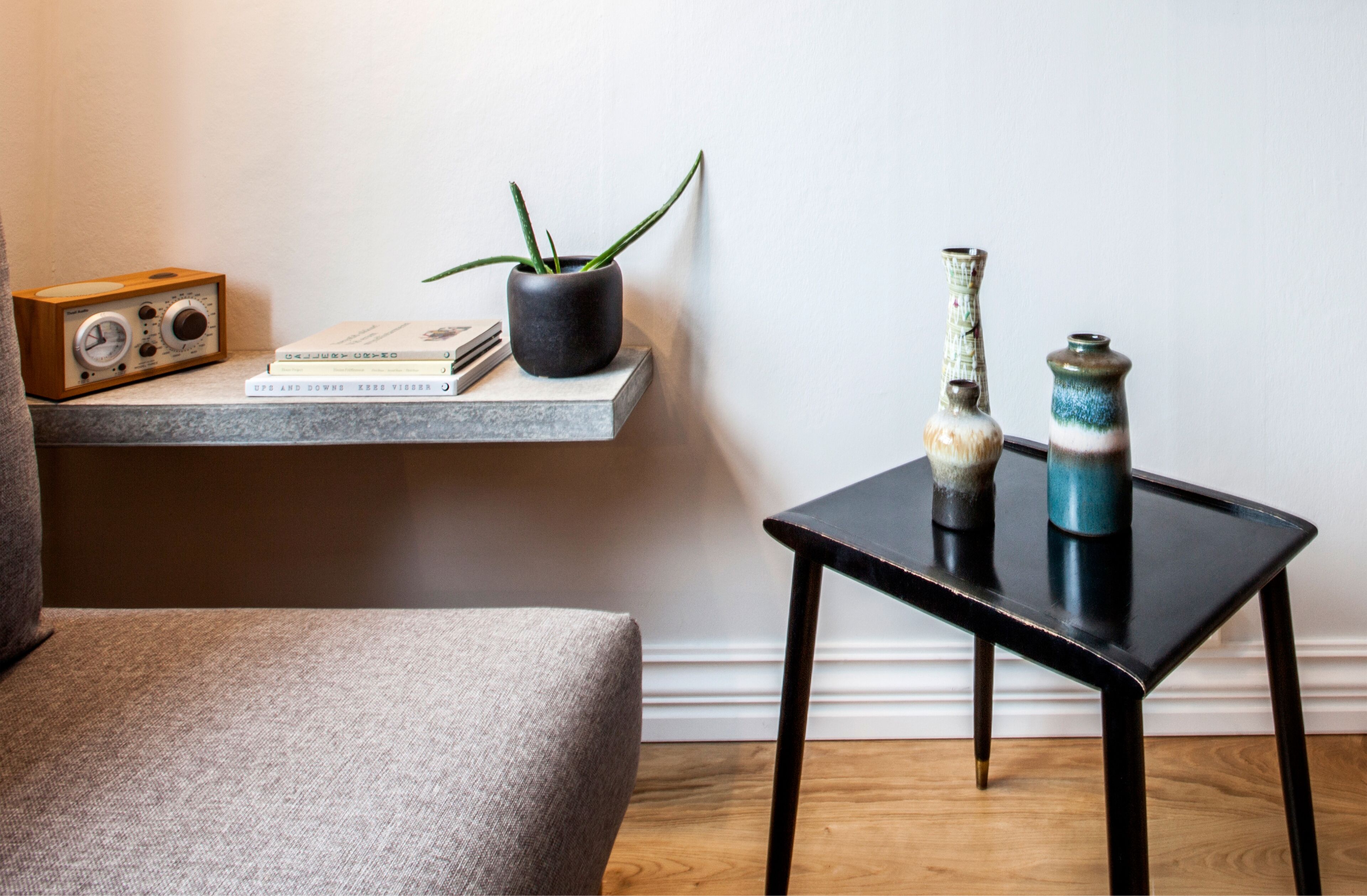 A room with a grey armchair, a small table with decorative vases, a radio, and a potted plant on a shelf.