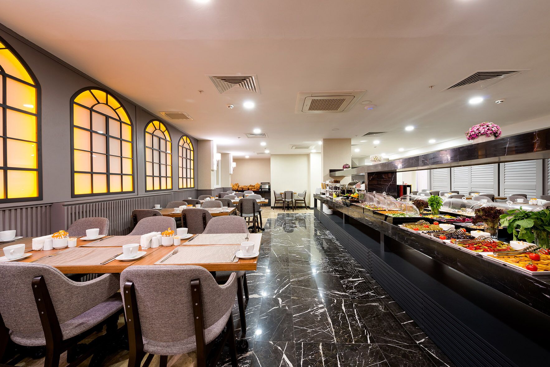 A buffet setup with a variety of food items, including salads and hot dishes, in a well-lit dining area with marble flooring and grey upholstered chairs.