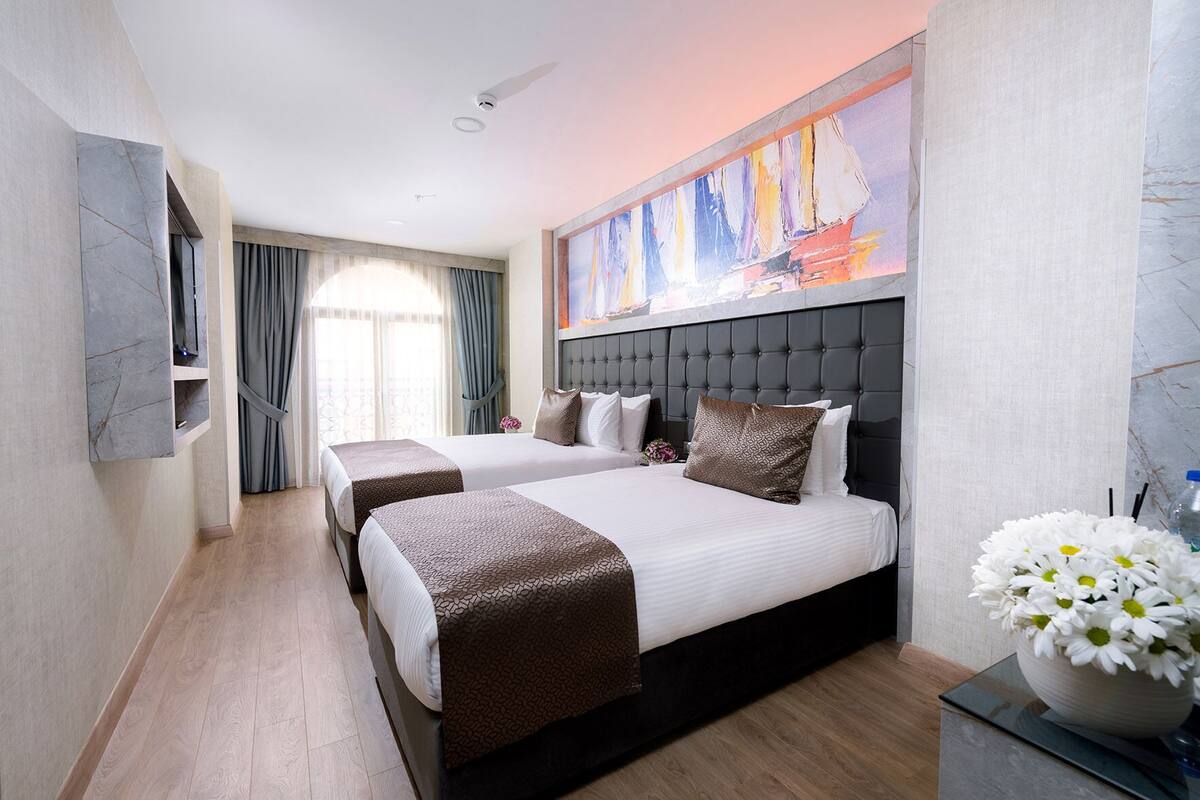 Quarto de hotel com duas camas, um quadro grande, um vaso de flores e vista para o exterior.