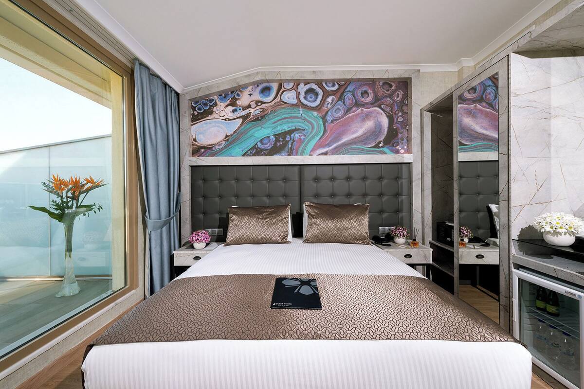 Quarto de hotel moderno com uma cama grande, cabeceira estofada, janela ampla com cortinas, um quadro na parede e um minibar.