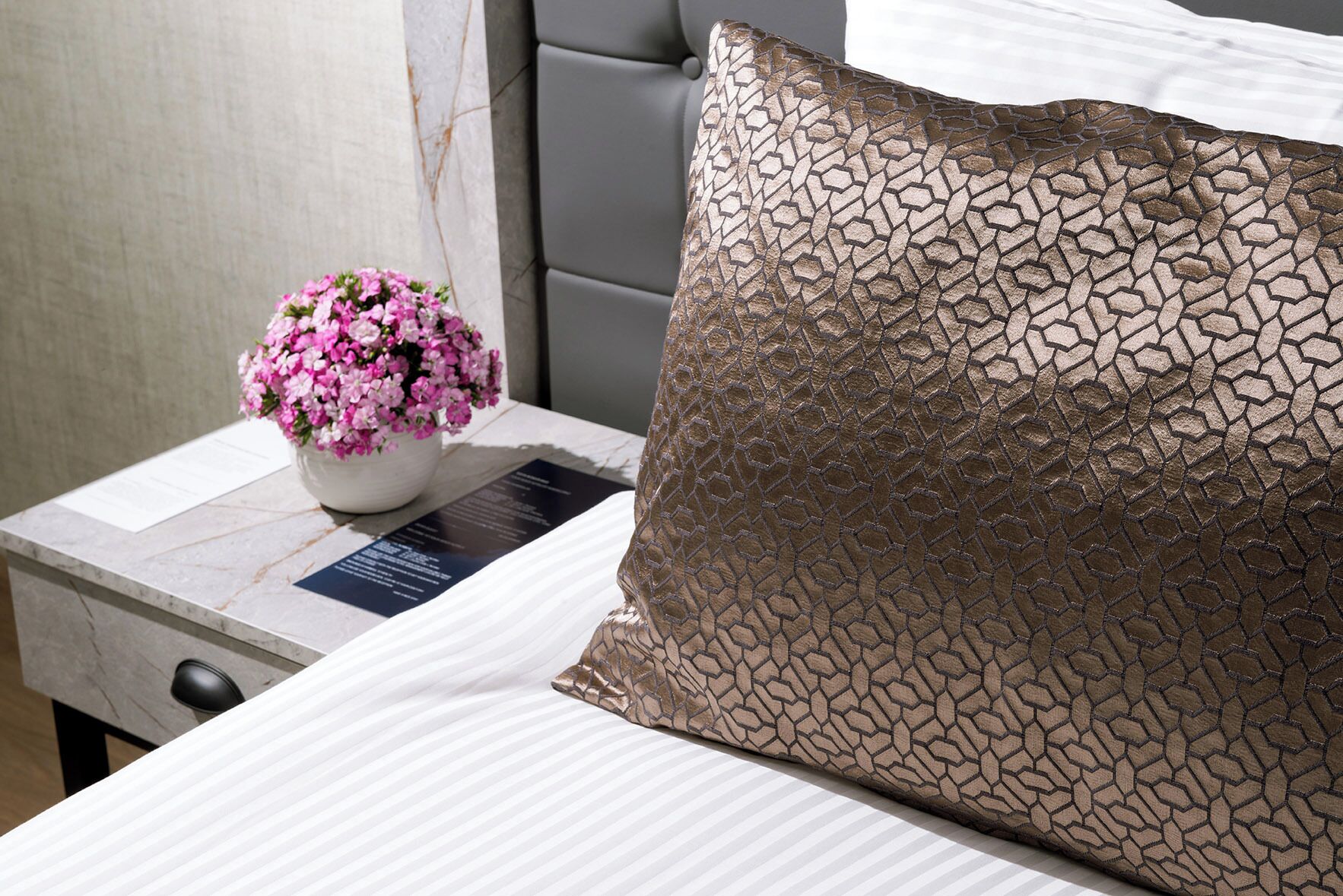 A neatly made bed with a patterned pillow and a small potted plant with pink flowers on a bedside table.