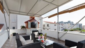 A covered outdoor seating area with wicker furniture, a glass table, and a brick oven. - Tihi Val Apartments (Split)