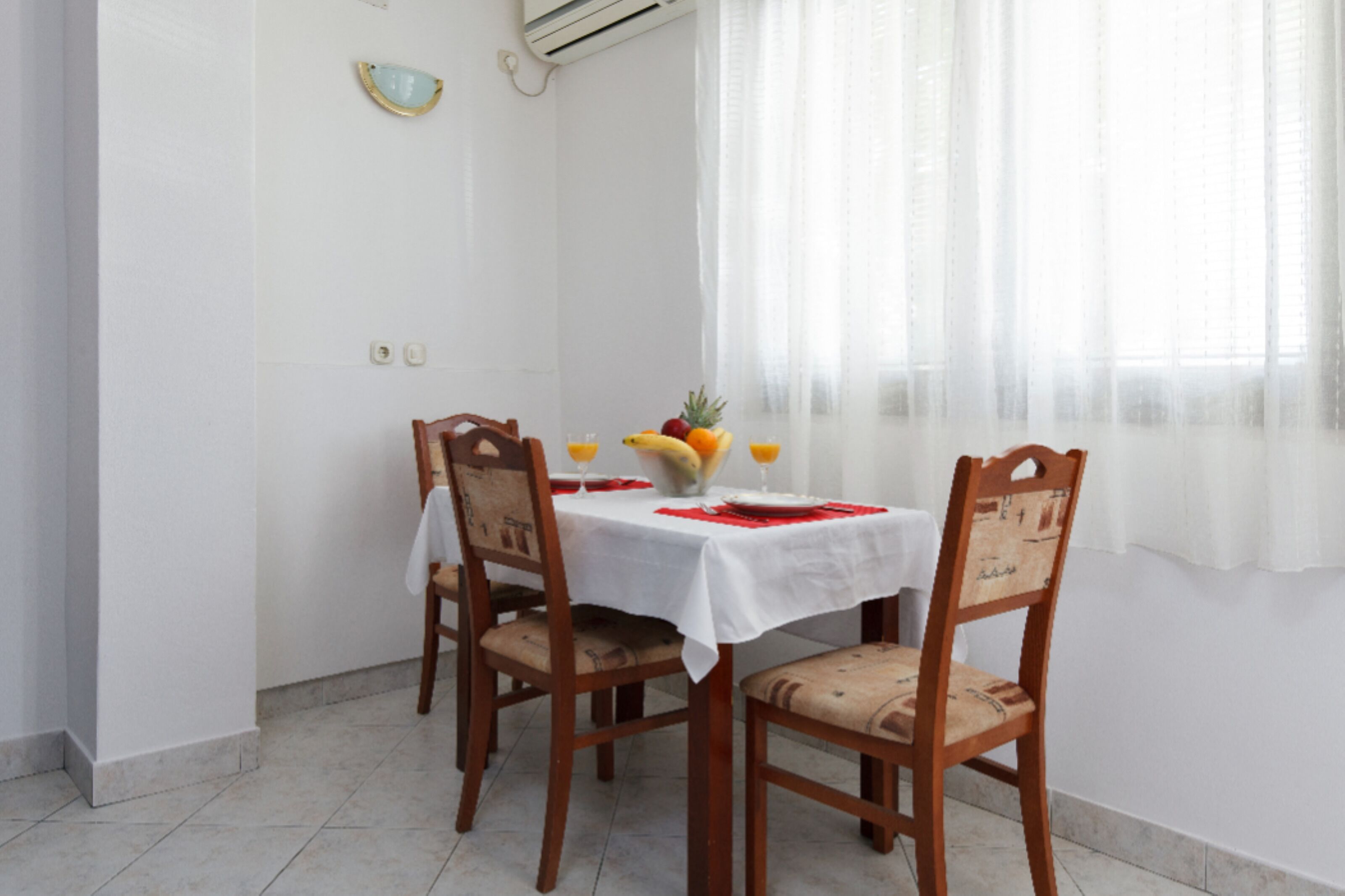A dining area with a white tablecloth, wooden chairs, and a fruit bowl with oranges and bananas.