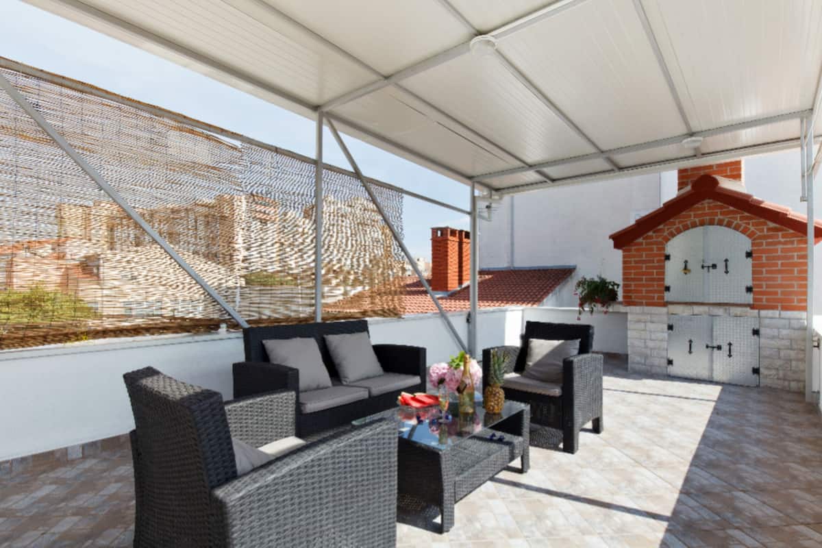 Une terrasse extérieure avec des meubles en osier, une table décorée de fleurs et une cheminée en briques.