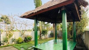 Una estructura metálica verde con techo de paja, posiblemente un porche o zona de estar al aire libre, rodeada de follaje y un sendero pavimentado.