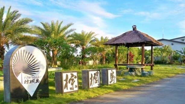Entrada del resort con un camino bordeado de palmeras, un pabellón de techo de paja y un cartel con el nombre del resort.