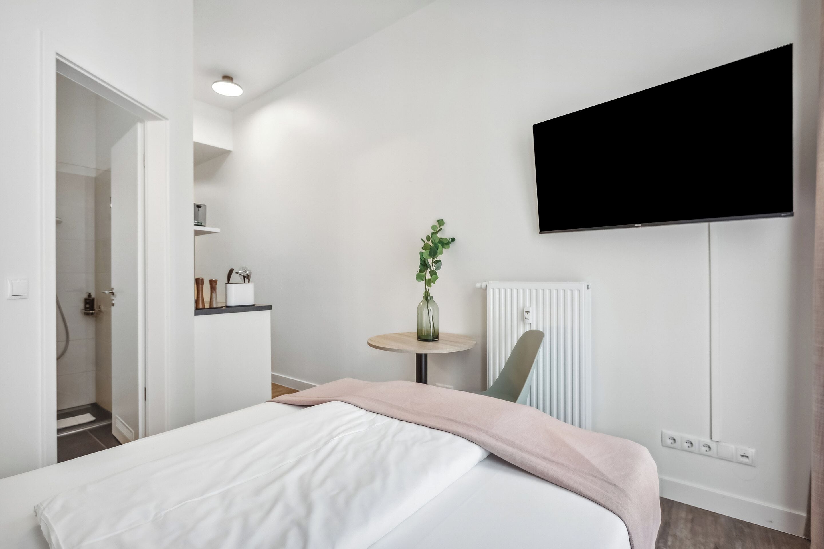 A modern hotel room with a bed, a small table with a vase, a chair, and a mounted TV.