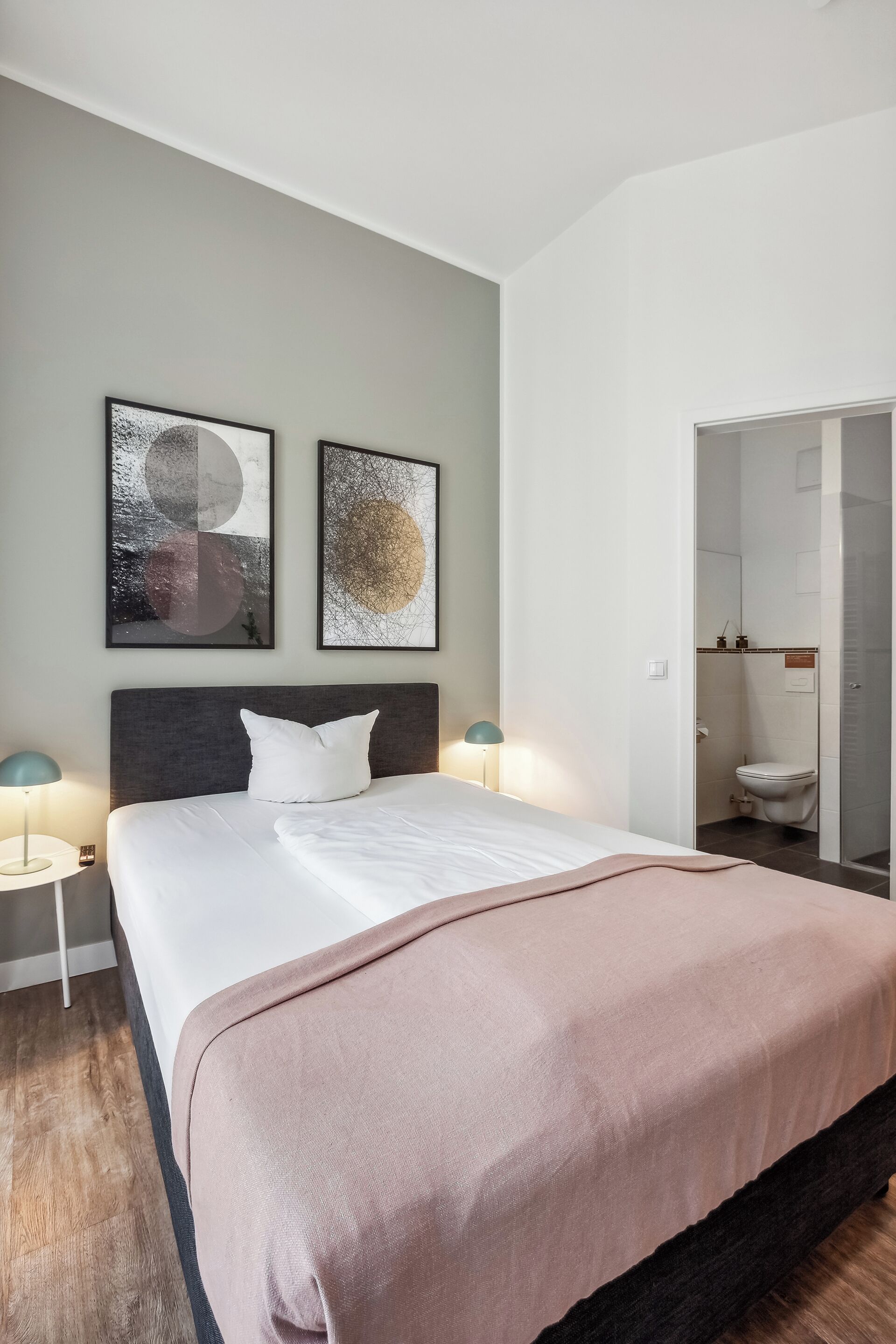 A modern hotel room with a large bed, two wall-mounted artworks, a bedside table with a lamp, and a bathroom visible through an open door.